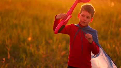 The-boy-in-the-costume-of-a-super-hero-running-in-a-red-cloak-laughing-at-sunset-in-summer-field-representing-that-he-was-the-pilot-of-the-plane-playing-with-a-model-airplane